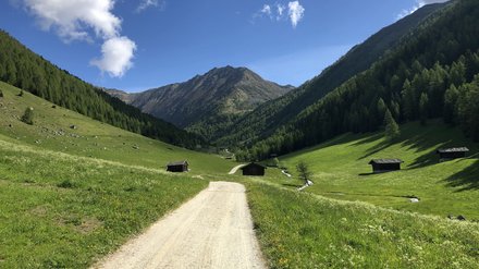 Schotterweg in den Bergen Schotterweg, der durch eine grüne Berglandschaft führt, umgeben von Wäldern und Hütten.
